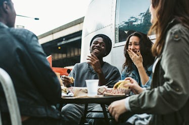 A group of friends laughs while sitting in an outdoor café and eating tacos.