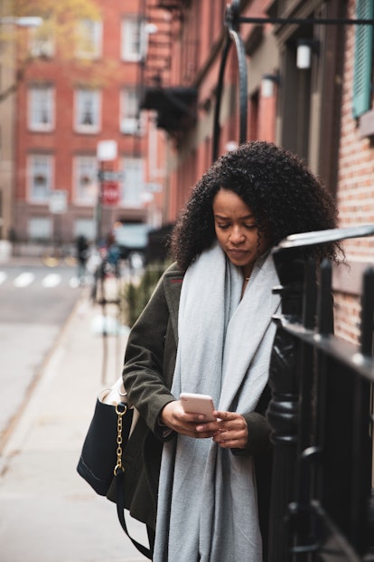A young woman quietly looks at her phone while leaning against a brick building in the city.