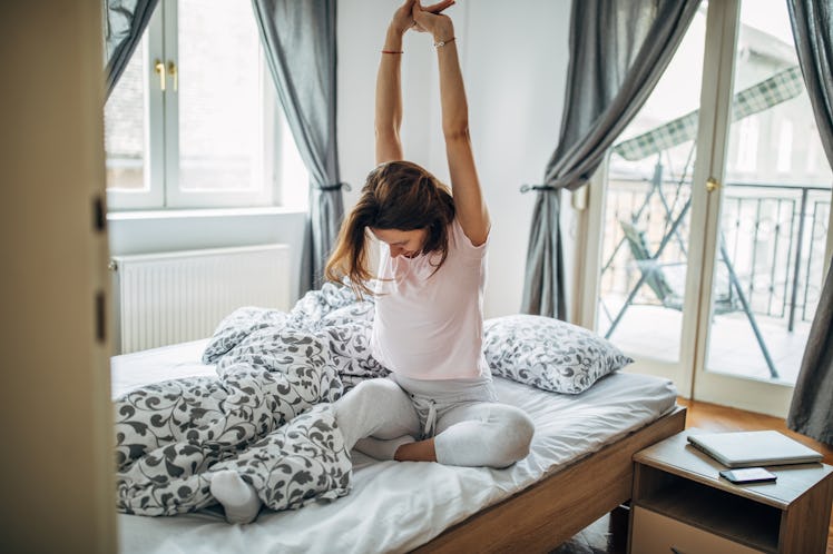 A young woman stretches in her bed in the morning while wearing comfortable clothes.