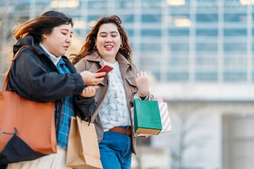 Two people walk together with shopping bags, with one person texting while her friend also looks at ...
