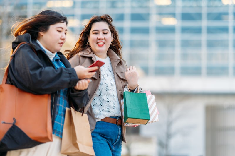 Two people walk together with shopping bags, with one person texting while her friend also looks at ...