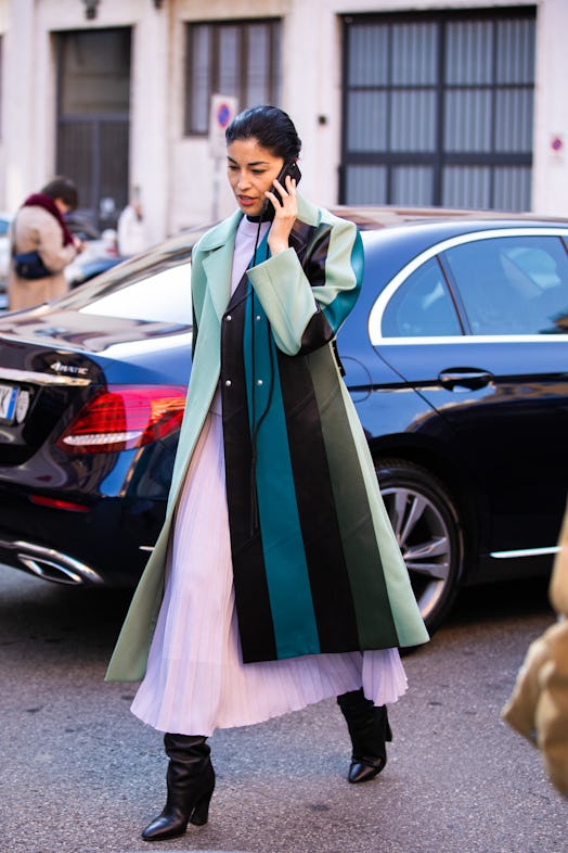 A woman walking in a long striped jacket with green, black and grey tones and a light pink dress und...