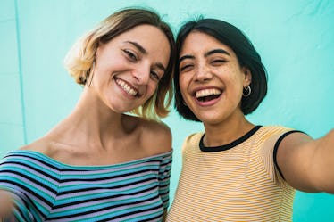 A happy couple stands in front of a brightly-colored wall and takes a selfie.