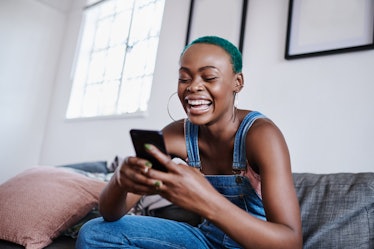 A woman laughs at her phone while sitting on a couch in her apartment.
