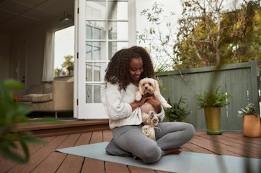 A young woman hangs out with her dog while sitting on a yoga mat on her deck.