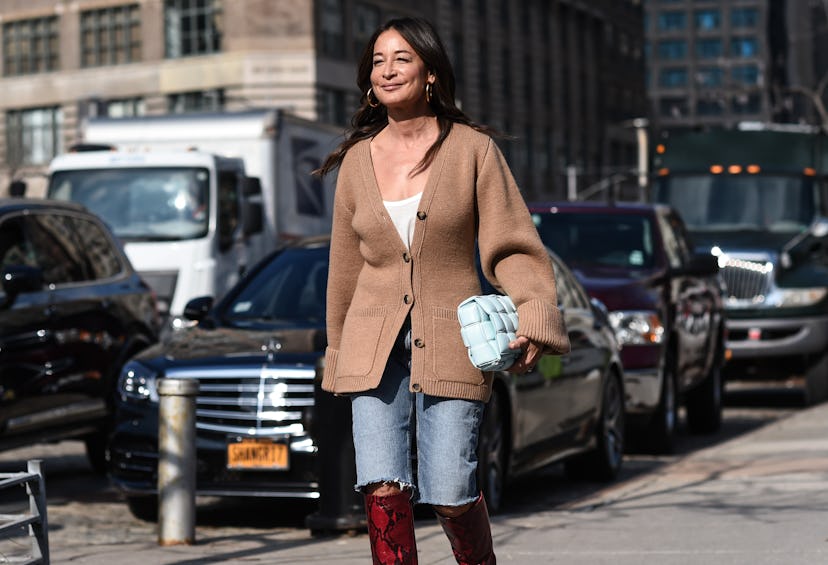 A woman walks the street in long denim shorts, a beige button-down sweater, and over-knee-high brown...