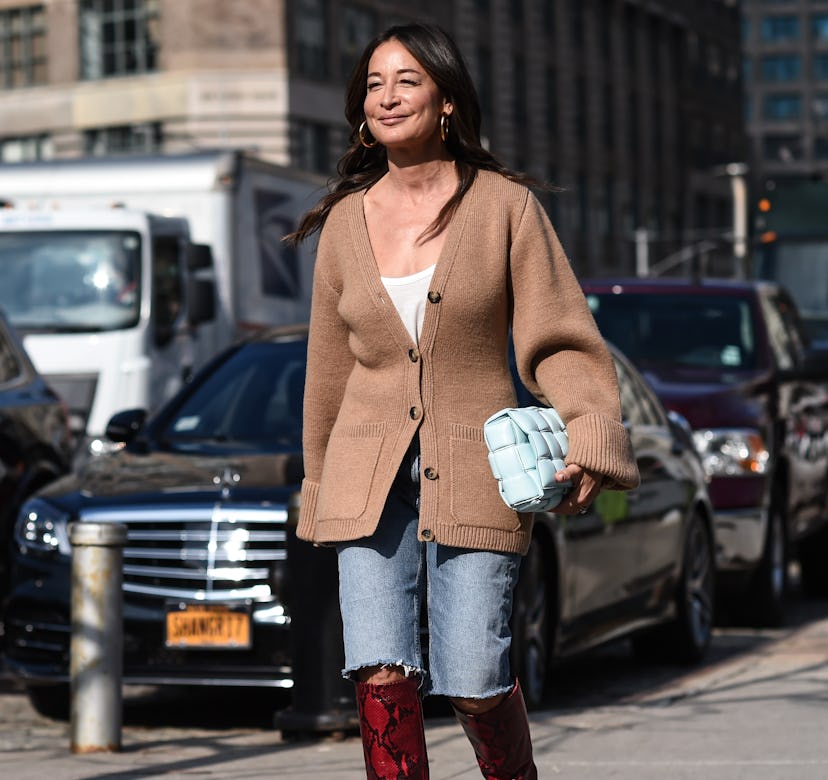 A woman walks the street in long denim shorts, a beige button-down sweater, and over-knee-high brown...