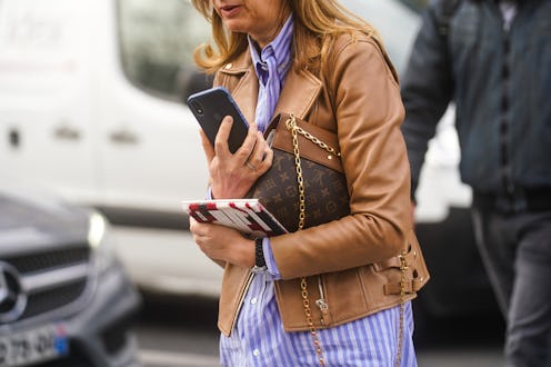A woman in a blue striped shirt and a caramel leather jacket, walking and carrying her phone and bro...