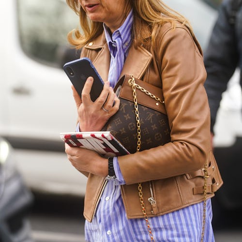 A woman in a blue striped shirt and a caramel leather jacket, walking and carrying her phone and bro...
