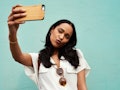 A young woman takes a selfie on her phone in front of a light blue wall.