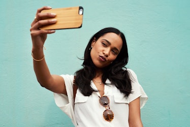 A young woman takes a selfie on her phone in front of a light blue wall.