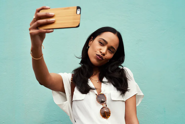 A young woman takes a selfie on her phone in front of a light blue wall.