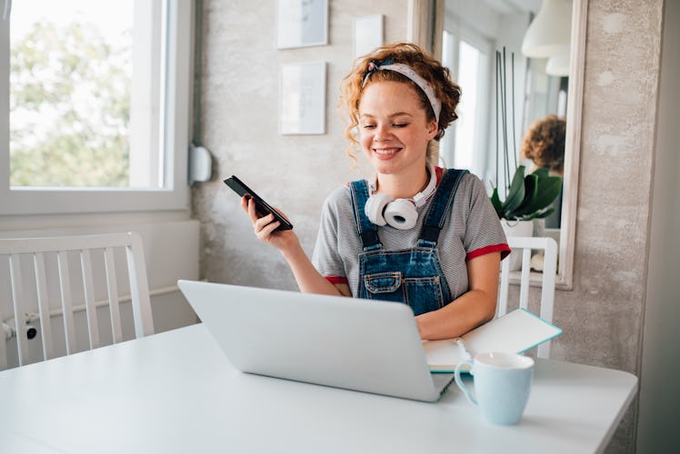 A young woman with red, curly hair sits at her laptop with her phone and a pair of headphones.