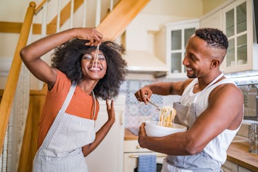 A young couple laughs and eats pasta in their kitchen.