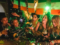 A group of friends dressed up in festive attire for St. Patrick's Day toast their drinks at a bar wh...