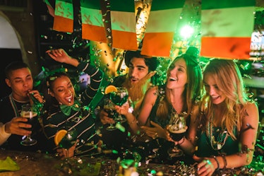 A group of friends dressed up in festive attire for St. Patrick's Day toast their drinks at a bar wh...
