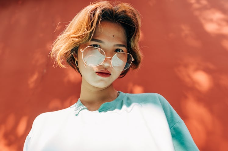 A young woman with glasses stands in a front of a bright coral wall on a sunny day.