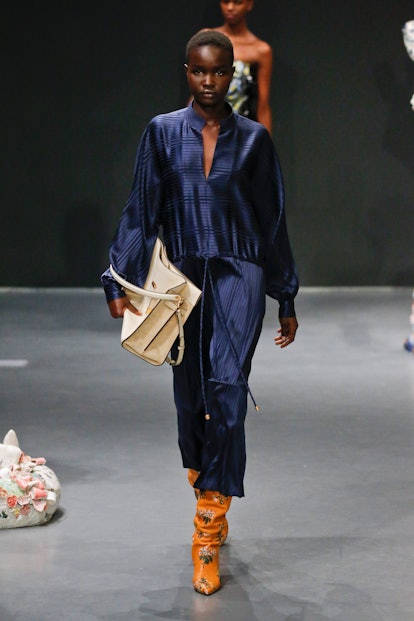 A model going down the runway in a blue outfit, orange boots and a white handbag all designed by ton...