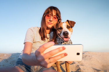 A young woman in pink sunglasses poses for a selfie on the beach with her dog.