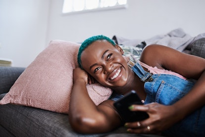 A young woman laughs while holding her phone and thinking on the couch.