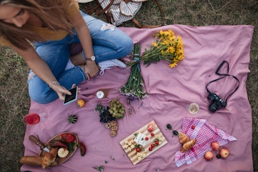 A young woman sits on a pink blanket in her backyard and has a picnic.