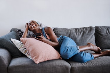 A young woman laughs while lounging on the couch and playing on her phone.