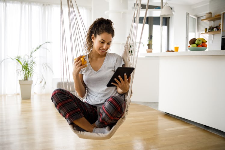 A young woman sits in a rope swing in her home while looking at her iPad.