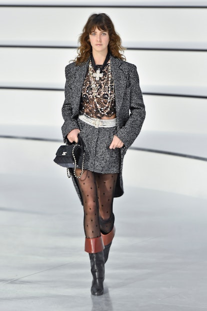 A model at the Chanel Fall 2020 Fall Show in a grey tweed blazer and skirt, a black mesh top, layere...