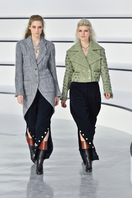 Two models on a runway at Chanel's 2020 Fall show wearing black skirts, one of them in a grey coat, ...