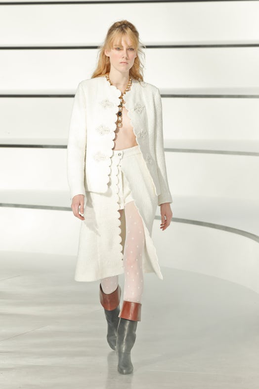 A model on a runway at Chanel's 2020 Fall Show in a white lace blazer and skort, and black-and-brown...