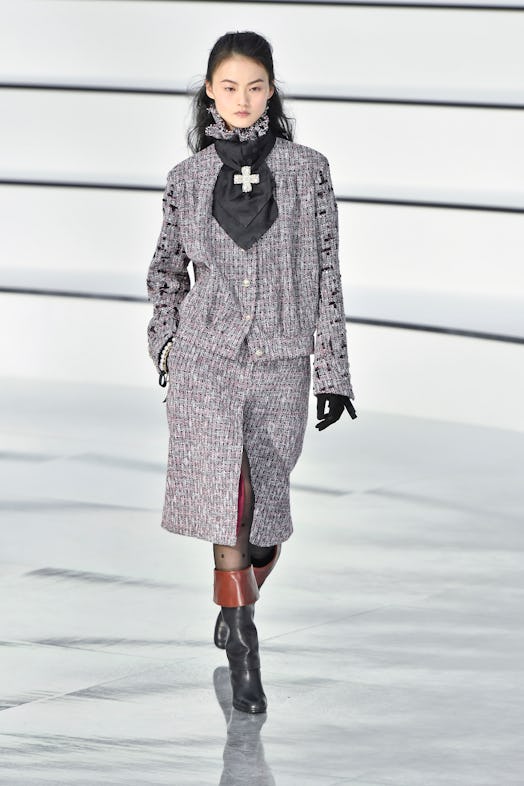 A model on a runway at Chanel's Fall 2020 in a grey tweed blazer and shirt, brown-black boots and a ...