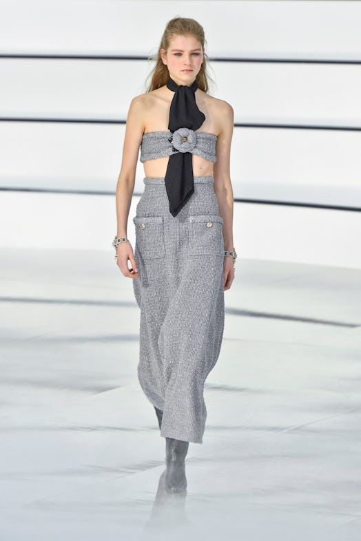 A model on a runway at Chanel's 2020 Fall show in a long grey tweed skirt and bandeau top, and a bla...