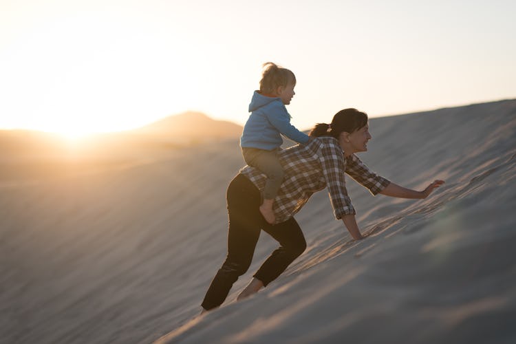 A mom carries her baby on her back in a sand dune.