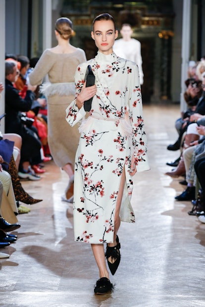 A model walking the runway in a white floral print dress paired with feather-trimmed flats