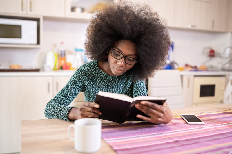 A young female with an afro hair, sitting at the kitchen table and reading a book about the history ...