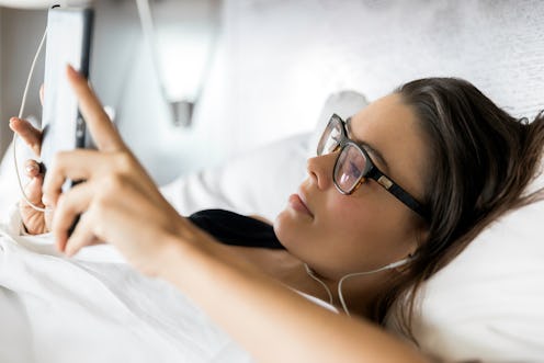 A woman wearing glasses looks at her phone in bed. Wearing your glasses instead of contacts is one w...