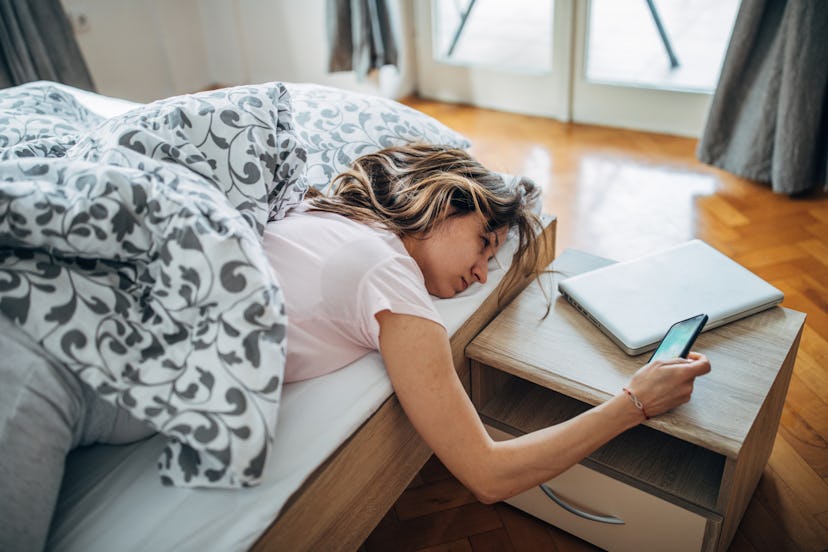 A person squints as she leans over her bed to check her phone. Blue light from your devices can prev...