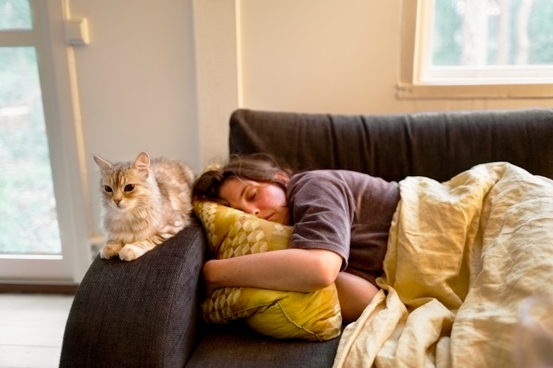 A woman sleeps on a couch with a cat perched on the armrest. Melatonin is a hormone that can help yo...