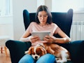 A young woman sits in a chair with her dog on her lap and uses her iPad.