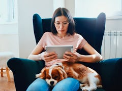 A young woman sits in a chair with her dog on her lap and uses her iPad.