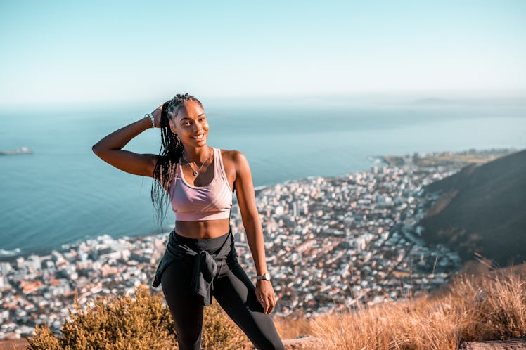 A smiling young woman in sportswear stands on a hilltop overlooking a coastal cityscape.