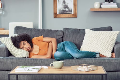A person lays on her couch, hugging herself and staring into the distance. Muscle aches are common w...