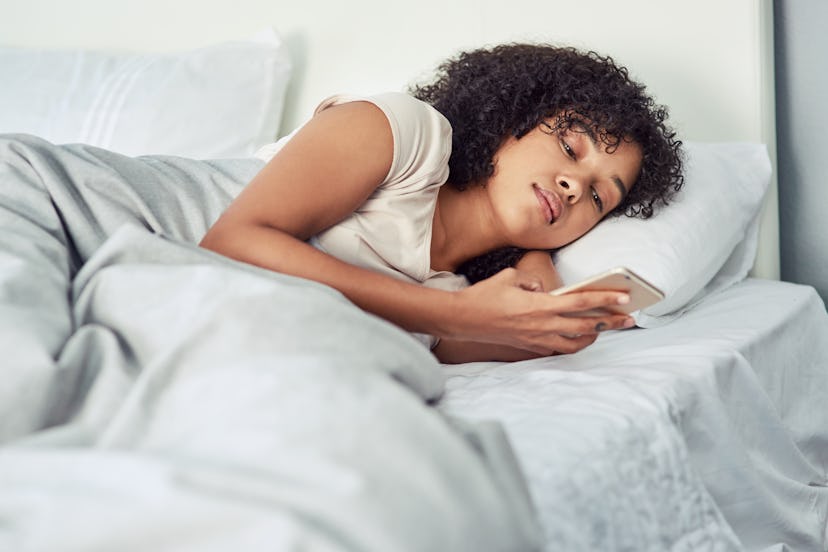 A woman looks at her phone in bed. Poor sleep can affect your cycle. 