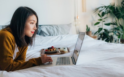 A young woman lays on her bed with her laptop and a brunch spread.