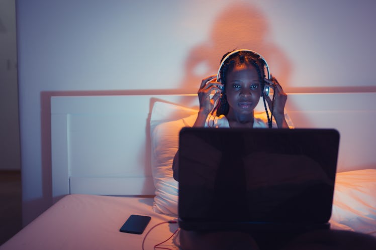 A young woman sits on her bed at night with her laptop and a pair of headphones on. 