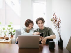 A young couple sits at a table in their apartment with a laptop and phone. 