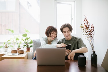 A young couple sits at a table in their apartment with a laptop and phone. 