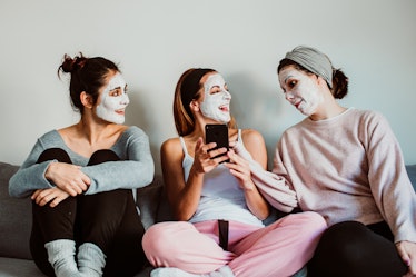 Three best friends hang out in sweatpants on a couch and do facials, while laughing at a phone.