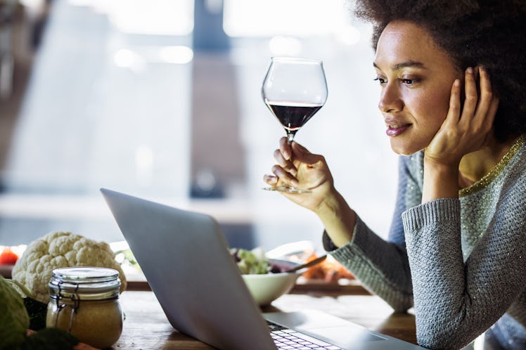A woman sits at a kitchen table with her laptop and a glass of red wine.