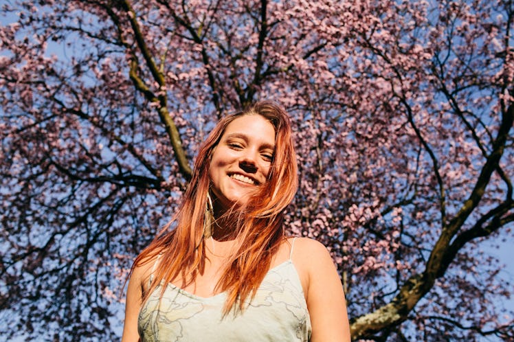 A young woman with red hair stands in the sunlight in front of a cherry blossom tree.